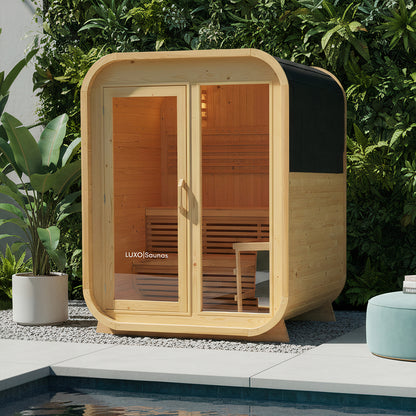 Waru 3 Person Traditional Finnish Outdoor Sauna in Natural, featuring a pod-shaped wooden design with a glass front, visible interior bench seating, and situated on a gravel patio beside a plunge pool.