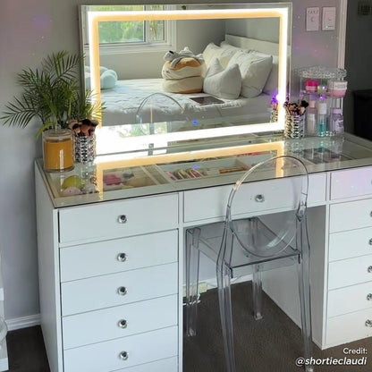 Adeline LED Mirror Storage Dressing Table in White features a sleek desk with glass-topped drawers, LED-lit mirror, and acrylic chair. Makeup brushes, skincare bottles, and a potted plant adorn the top.