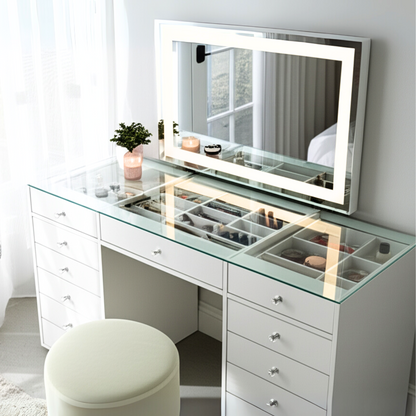 Adeline LED Mirror Storage Dressing Table in White features a glass countertop revealing organized cosmetics. It includes a large LED-lit mirror, multiple drawers, a potted plant, and a round stool.