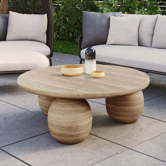 The Alessia 100cm Round Concrete Coffee Table in Beige features a round tabletop with three spherical bases, complemented by two wooden bowls and a two-tone decorative vessel, set in an outdoor seating area.