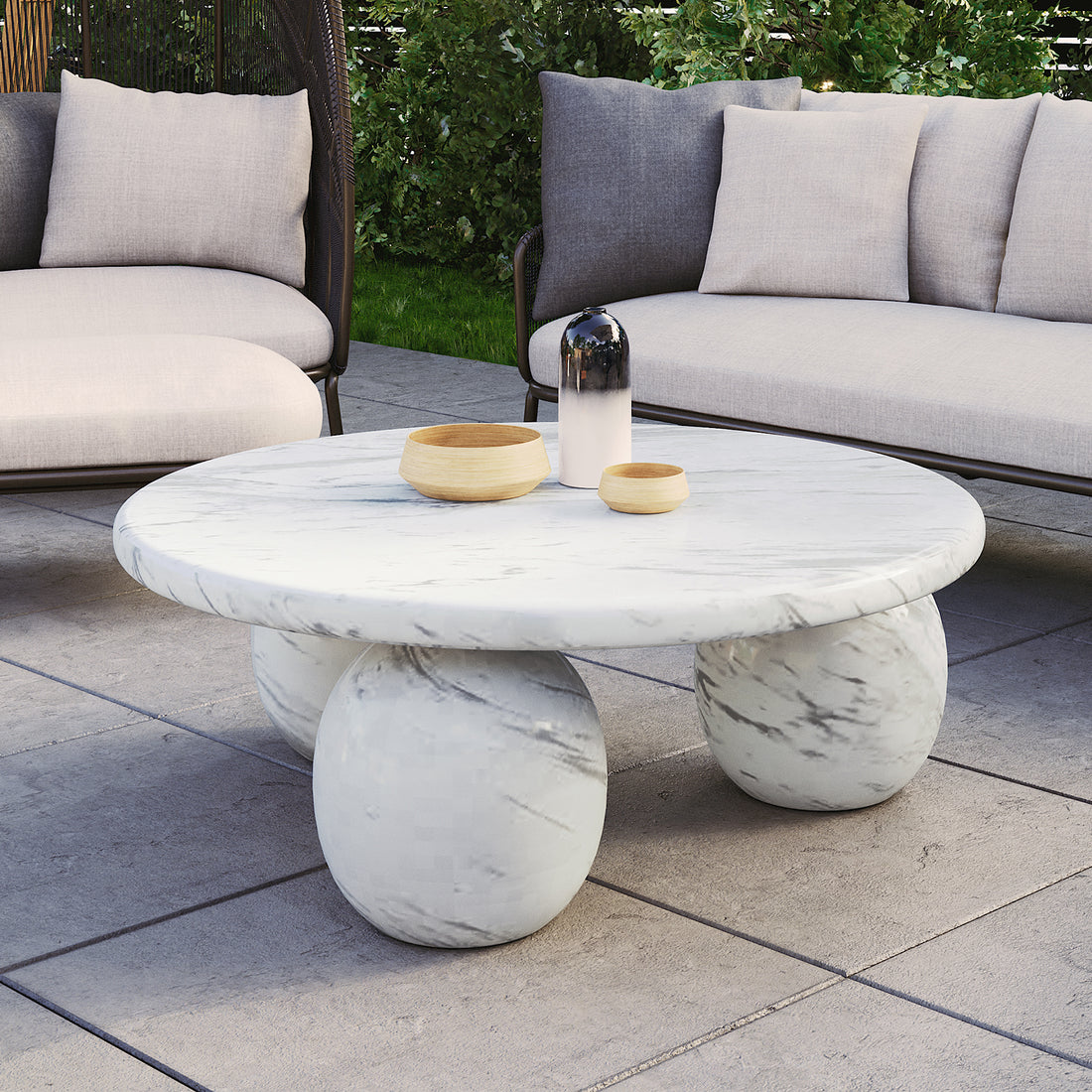 Alessia 100cm Round Concrete Coffee Table in White Marble features a marble top with three spherical marble legs, topped with two wooden bowls and a gradient decorative vessel, set on a patio.