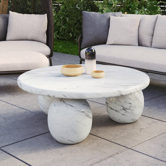 Alessia 100cm Round Concrete Coffee Table in White Marble features a marble top with three spherical marble legs, topped with two wooden bowls and a gradient decorative vessel, set on a patio.