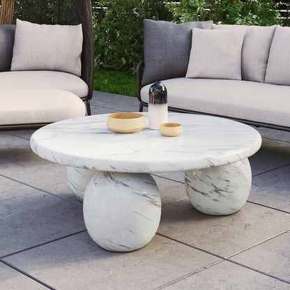 Alessia 100cm Round Concrete Coffee Table in White Marble features a marble top with three spherical marble legs, topped with two wooden bowls and a gradient decorative vessel, set on a patio.