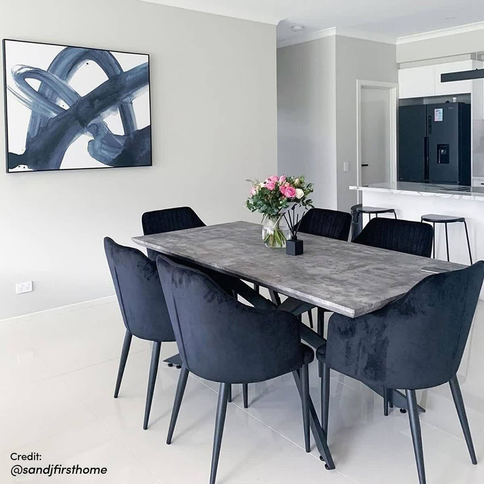 Alfred 180cm 6 Seater Pedestal Dining Table in Concrete features a concrete-look top with sleek black metal legs, surrounded by six black upholstered chairs, complemented by a vase of blooms and a reed diffuser.