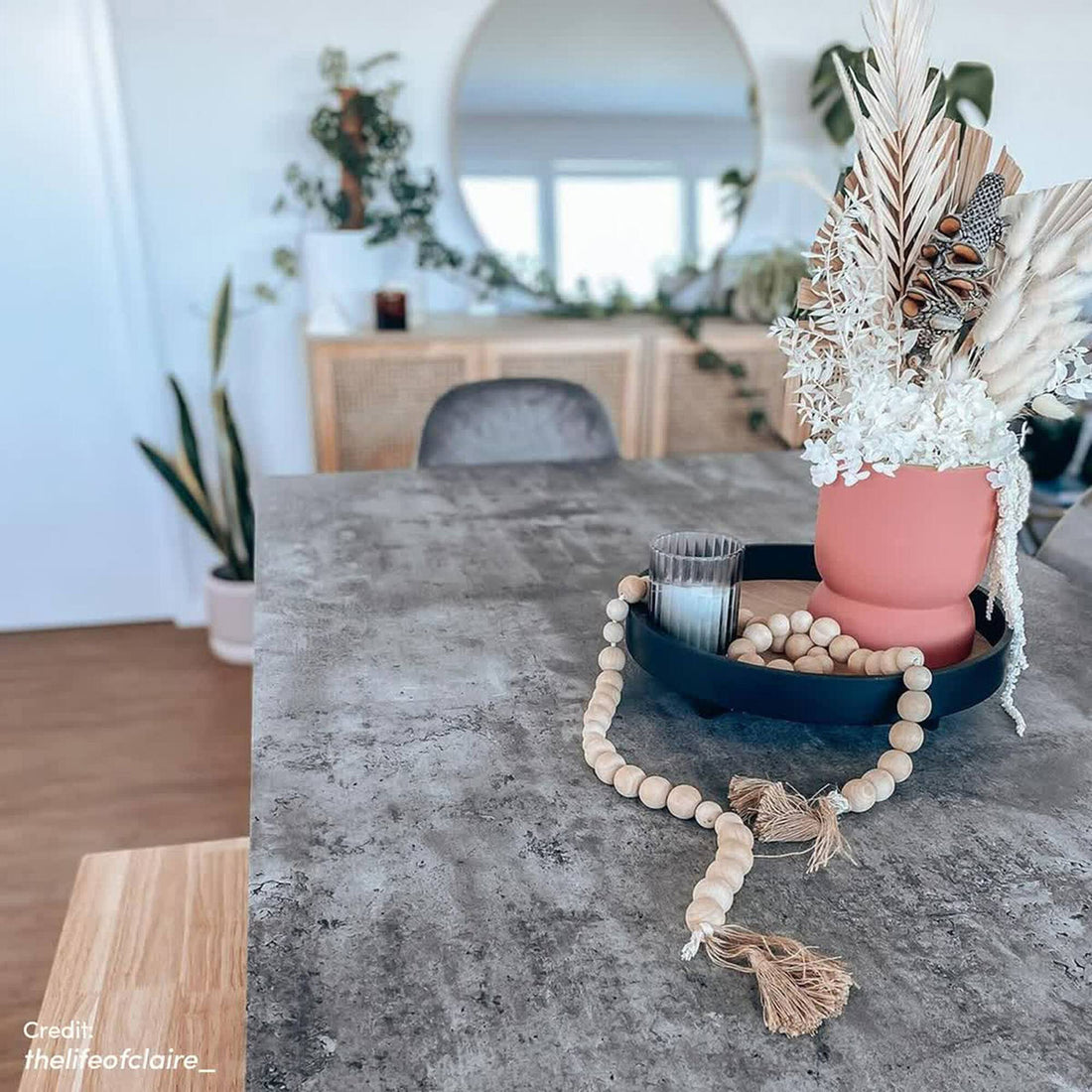 Alfred 180cm 6 Seater Pedestal Dining Table in Concrete is showcased with a grey countertop, featuring a black tray, pink-coral vase, ribbed glass, and natural wood beads, in a modern, boho interior.