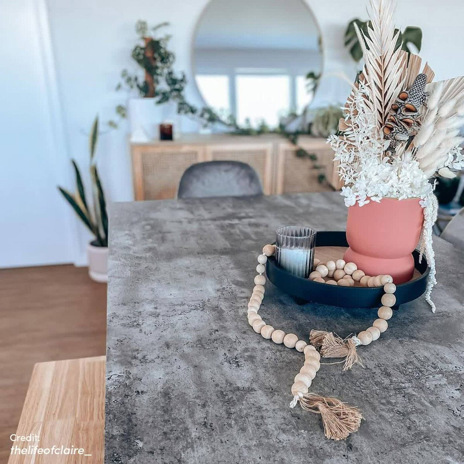 Alfred 180cm 6 Seater Pedestal Dining Table in Concrete is showcased with a grey countertop, featuring a black tray, pink-coral vase, ribbed glass, and natural wood beads, in a modern, boho interior.