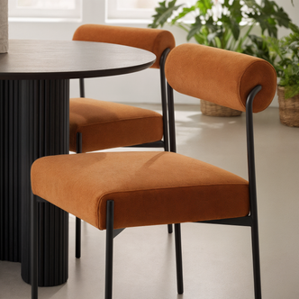 Alona Modern Velvet Dining Chair Set of 2 in Terracotta features rust-orange upholstered seats, cylindrical backrests, and slim black legs, paired with a round black fluted table for a chic dining arrangement.