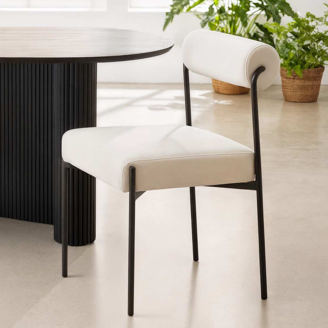 Alona Modern Velvet Dining Chair Set of 2 in Cream White, featuring a slim black metal frame, padded white seat, and cylindrical back cushion, with a round table edge visible in a plant-filled room.