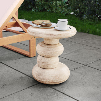 Amalee 40cm Round Concrete Side Table in Travertine, featuring a sculptural base of stacked stone-like discs, topped with a teacup on a saucer, set on a patio with lounge chair nearby.