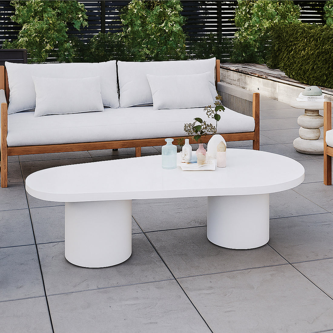 Amarosa Curved Indoor/Outdoor Concrete Coffee Table White with a sculptural design, featuring two cylindrical pedestal legs, topped with decorative vases and foliage, set in a modern outdoor seating area.