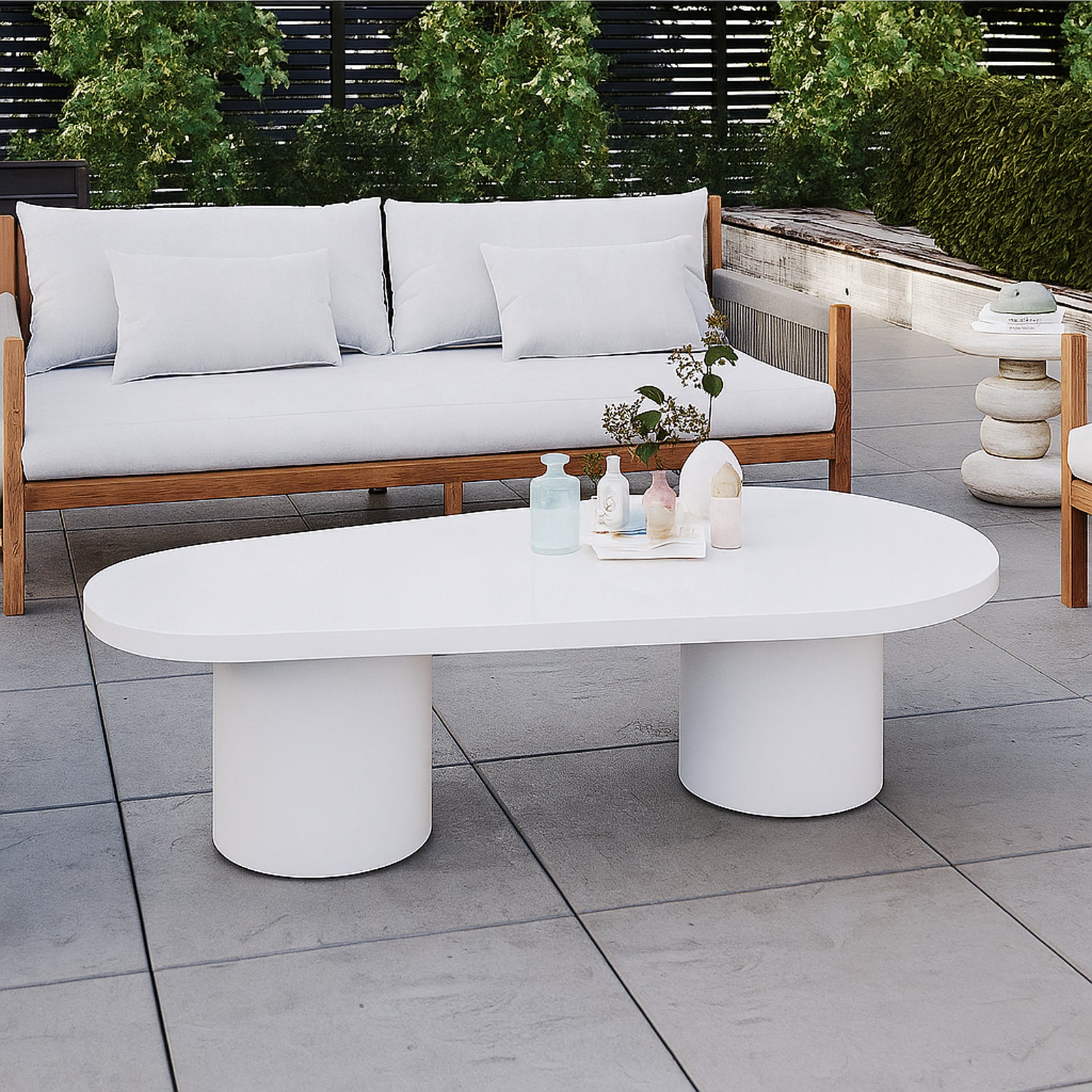 Amarosa Curved Indoor/Outdoor Concrete Coffee Table White with a sculptural design, featuring two cylindrical pedestal legs, topped with decorative vases and foliage, set in a modern outdoor seating area.