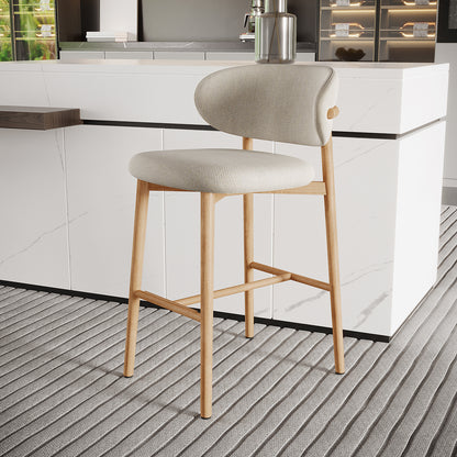 The Audrey Curved Linen Barstool Set of 2 in Beige features a light wood frame, slender legs, and a softly upholstered light-gray seat, complementing a modern kitchen with a marble-style counter.