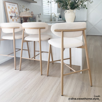 Audrey Curved Boucle Barstool Set of 2 in Cream White features upholstered seats with light wooden legs, placed near a kitchen counter with a vase and coffee station. Perfect for modern spaces.