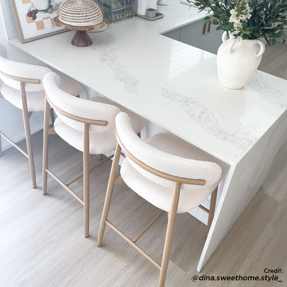 Audrey Curved Boucle Barstool Set of 2 in Cream White, featuring light wood frames and plush boucle upholstery, lines a modern, minimalist kitchen island with a sleek waterfall-edge quartz countertop.