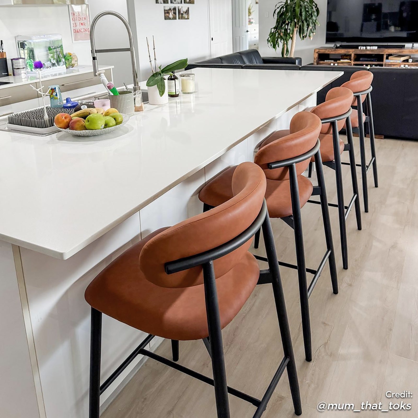 Audrey Curved PU Leather Barstool Set of 2 in Tan, featuring plush tan-brown seats with black metal legs, positioned around a sleek white island in a modern open-plan kitchen and living area.