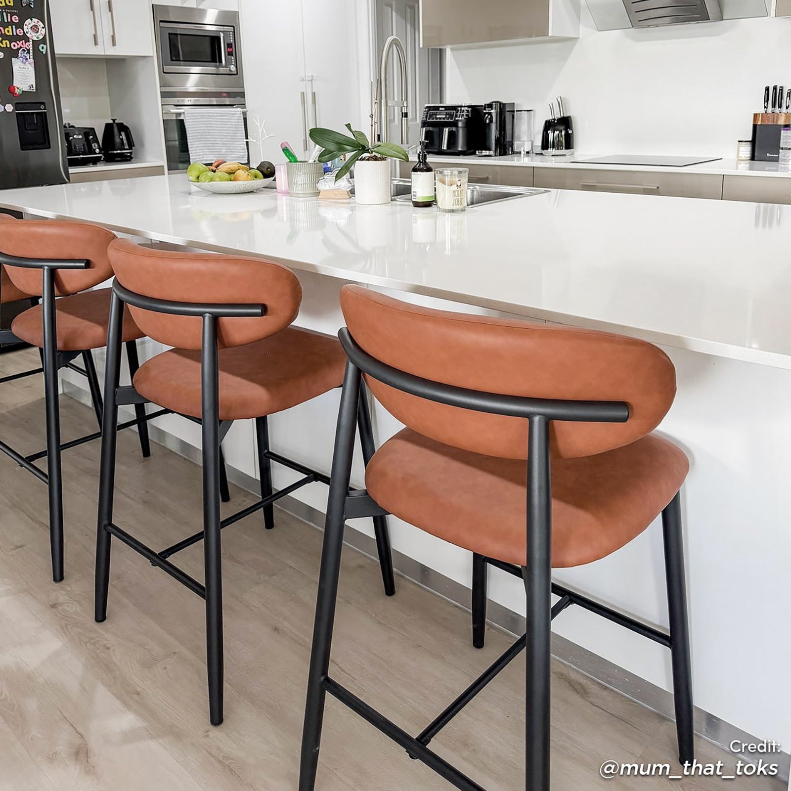 Audrey Curved PU Leather Barstool Set of 2 in Tan features rust-brown padded seats with black metal frames, placed alongside a modern kitchen island, enhancing contemporary elegance and comfort.