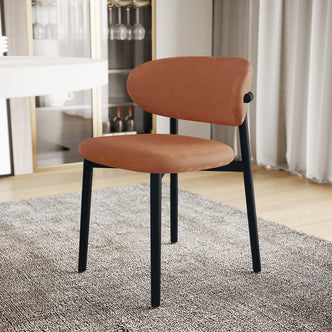 Audrey Curved Leather Dining Chair Set of 2 in Tan features rounded terracotta upholstery, slim black metal legs, on a gray rug, with a nearby white table and glass-fronted cabinet backdrop.