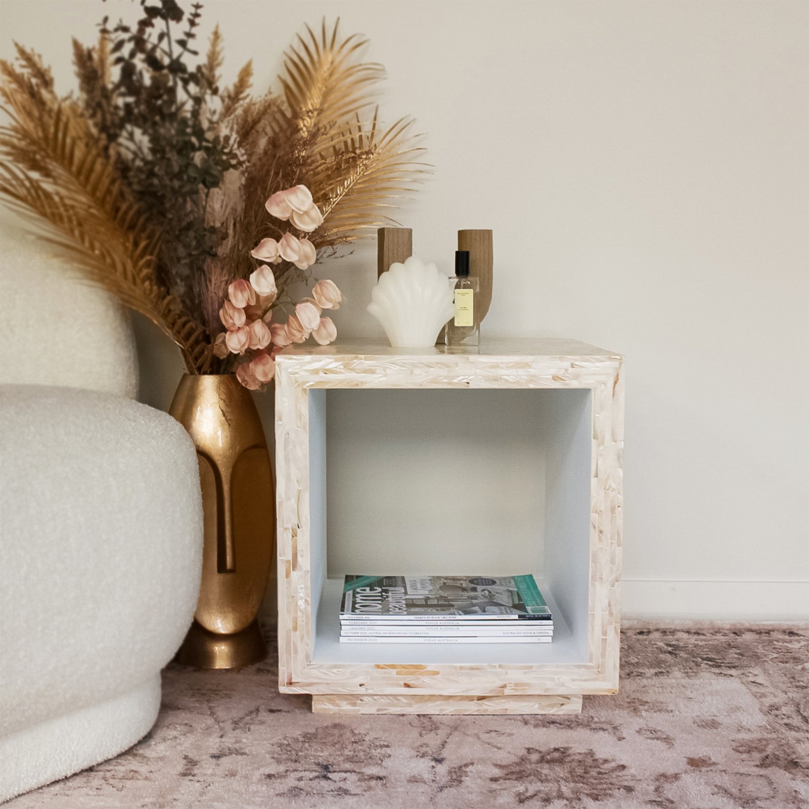 Aurora 42cm Mother of Pearl Inlay Side Table - Stripe Beige with a sculptural shell candle, glass bottle, and empty wooden candle holders on top; magazines inside the cubby, near a boucle armchair.