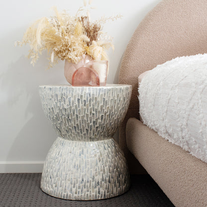 Baylor 40cm Mother of Pearl Inlay Side Table, Pale Blue, showcases a two-tiered hourglass form with a mosaic pattern, adorned with a pink ceramic vase, glass candle holder, and decorative arch sculpture.