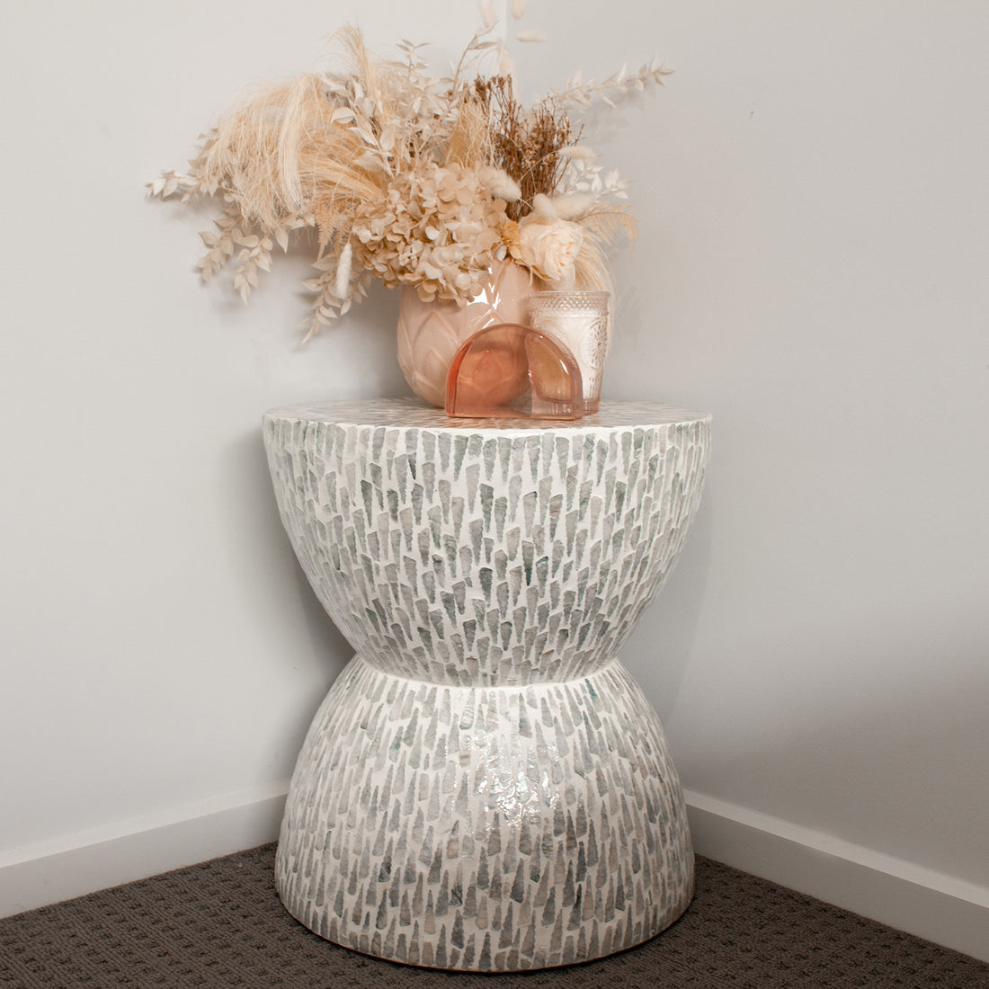 Baylor 40cm Mother of Pearl Inlay Coffee Table Pale Blue, featuring a unique hourglass shape with mosaic pattern, topped with a pink vase of dried botanicals and decorative glass items.