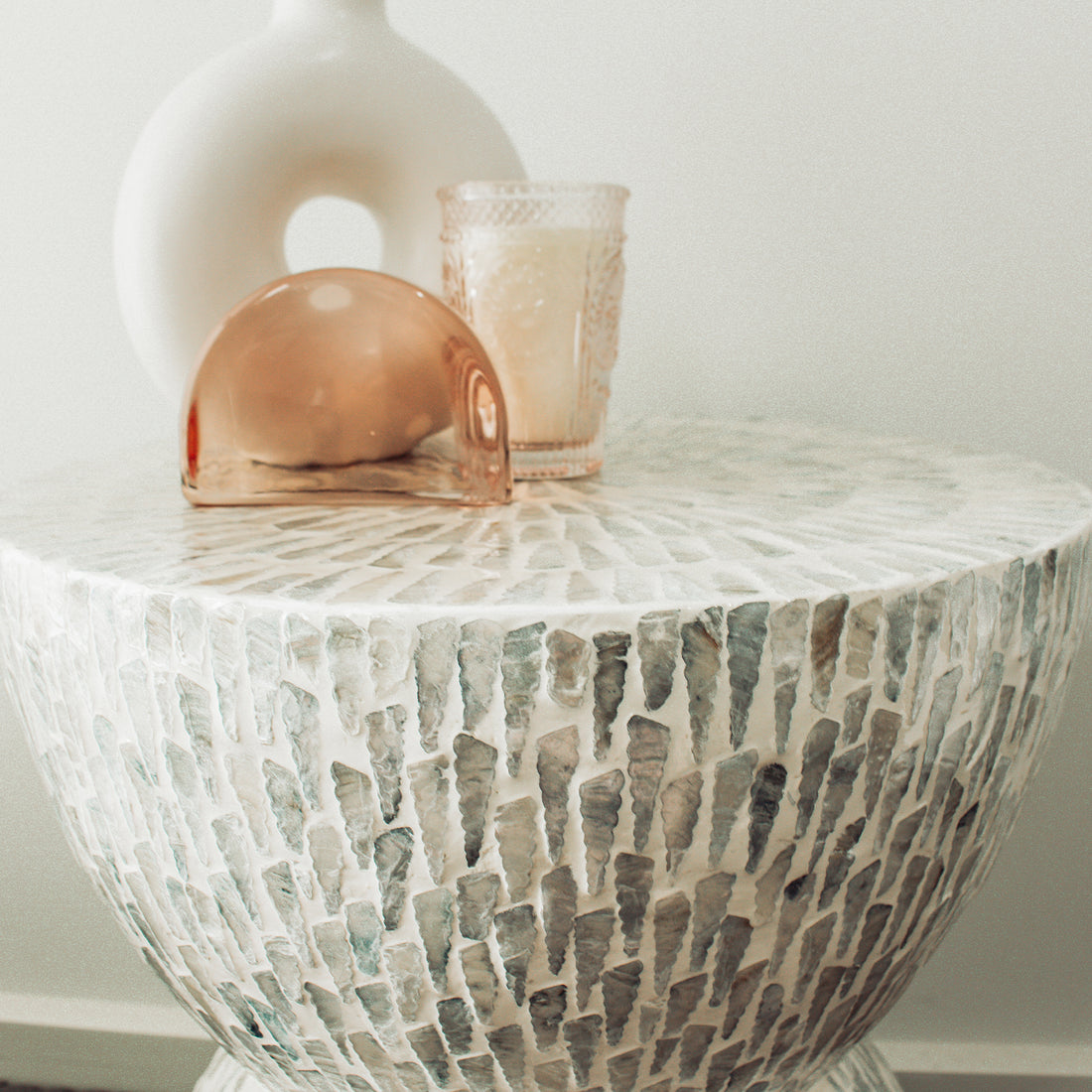 Baylor 40cm Mother of Pearl Inlay Side Table with stone or shell tiles, featuring a ceramic donut vase, textured tumbler with creamy wax, and a translucent peach-pink glass sculpture on top.