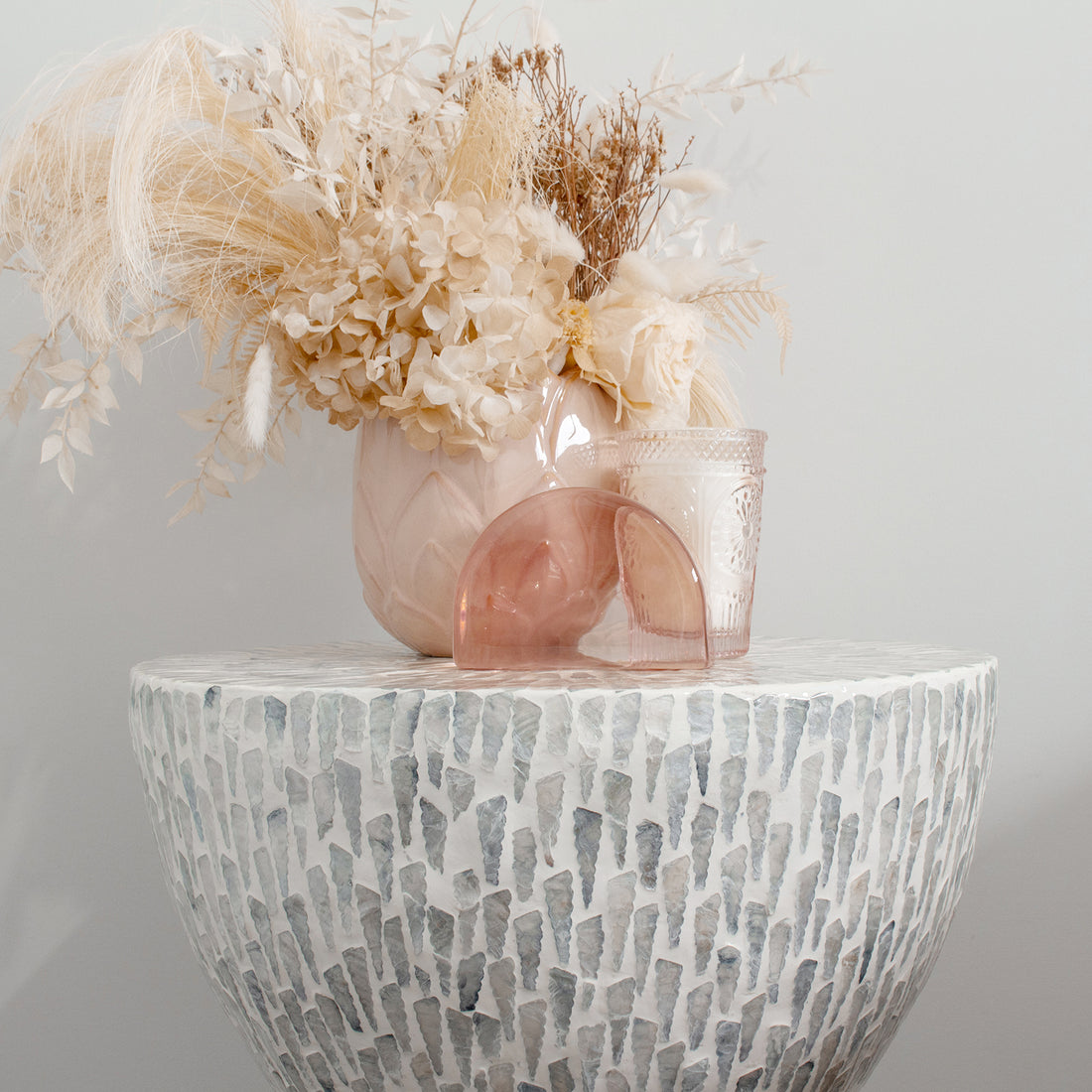 Baylor 40cm Mother of Pearl Inlay Coffee Table Pale Blue with a mosaic surface, blush vase of dried blooms, pink glass votive, and arch sculpture creating a minimalist, neutral-toned vignette.