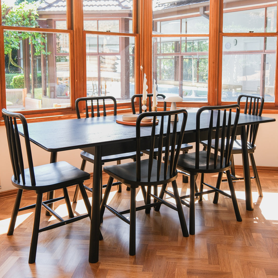 Angled view of the Luxo Living Bijan extendable black oak dining table with six matching Leigh spindle-back chairs, set in a bright dining room with large windows overlooking greenery, showcasing elegant decor.