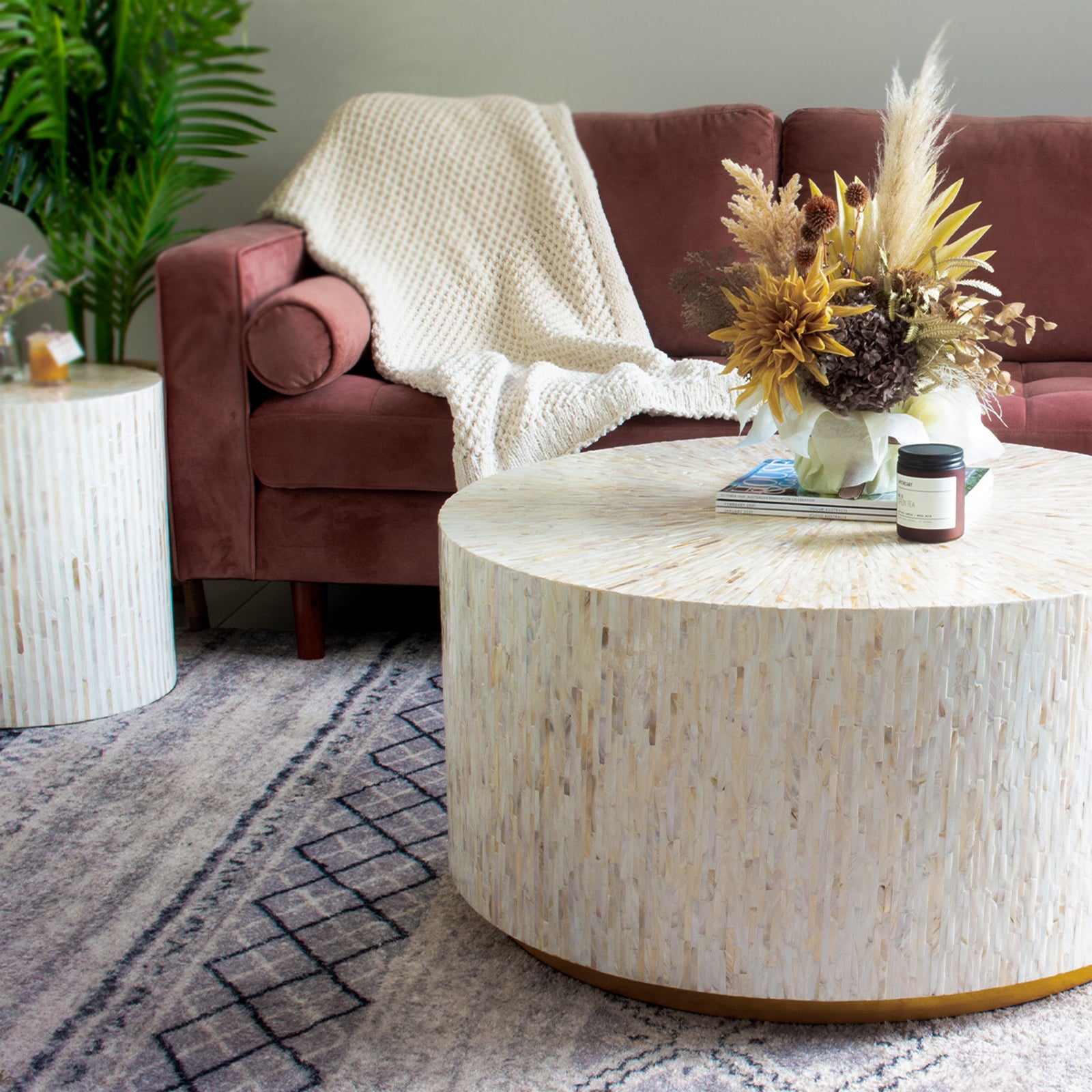 Bisera 91cm Mother of Pearl Inlay Coffee Table with intricate mother-of-pearl mosaic top, displayed in a cozy indoor setting on a couch, alongside a brown jar and flowers on a table.