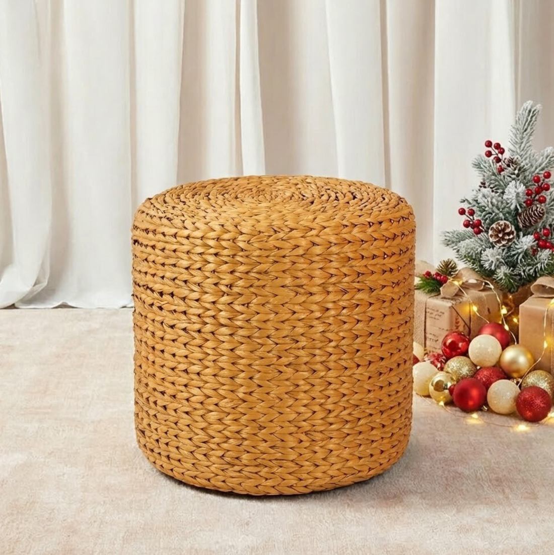 Carla 40cm Water Hyacinth Pouffe Ottoman in Honey, woven from natural fibers, sits on a light rug, with festive decor featuring evergreen sprigs, gifts, and Christmas baubles in the background.