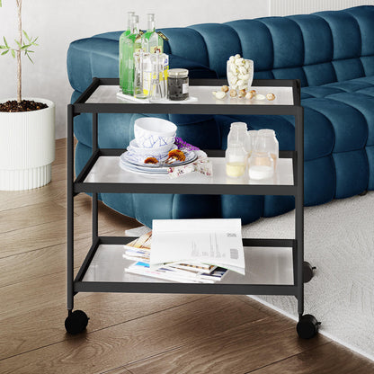 Chelsea 3-Tier Fluted Glass Drink Cart Bar Trolley in Black, featuring three fluted glass shelves displaying spirits, mixing beakers, bowls, and magazines, positioned on casters in a modern living-room corner.