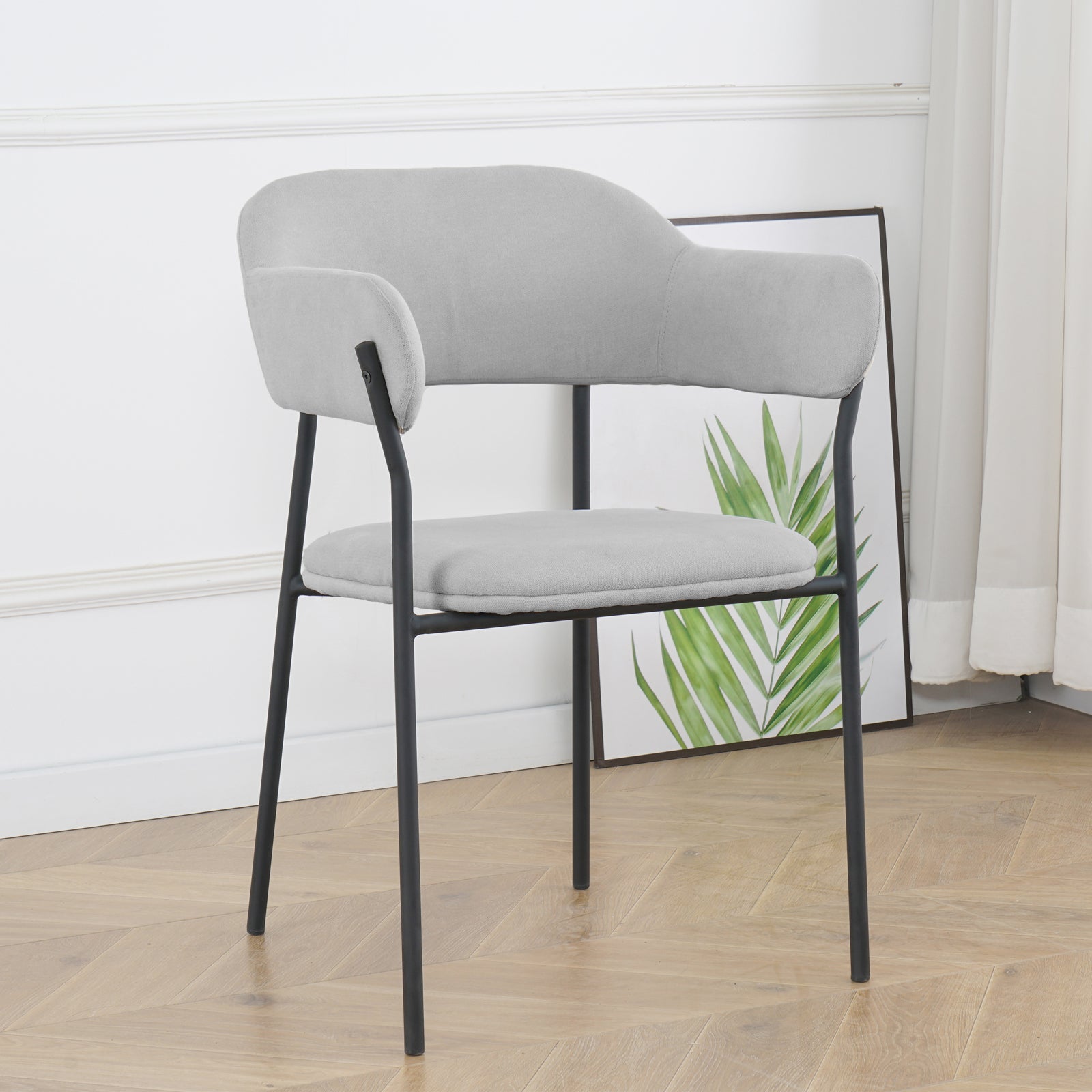 Clavin Fabric Dining Chair Set of 2 in Light Grey, features a modern design with padded upholstery, curved backrest, and slender black metal legs. Positioned in a minimalist setting with a palm print backdrop.