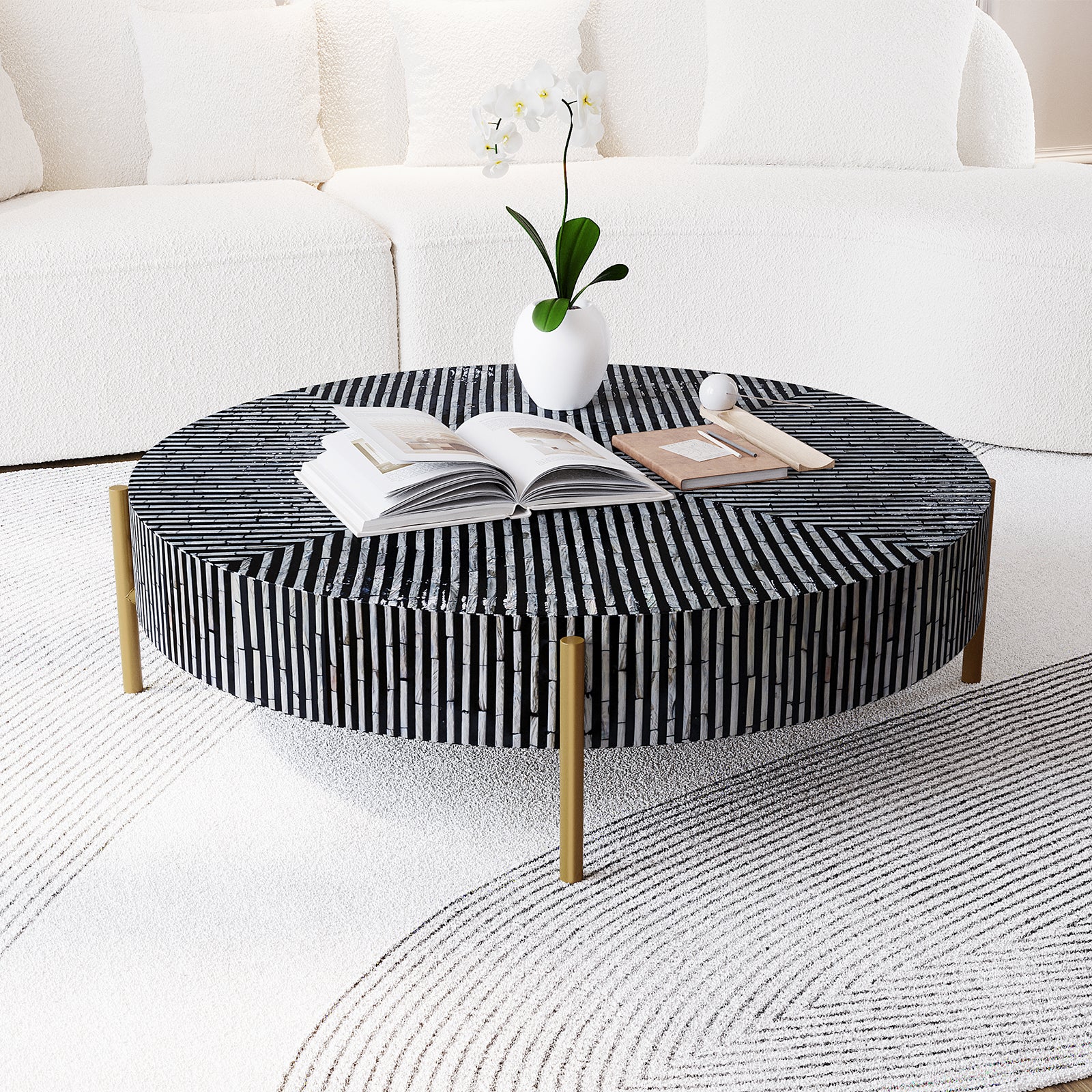 Cradily Mother of Pearl Inlay Coffee Table in Ivory & Black with mosaic pattern tabletop, brass legs, open book, notebook with pen, decorative sphere, orchid vase, plush sofa, and detailed rug backdrop.