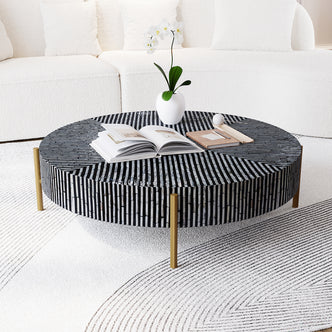 Cradily Mother of Pearl Inlay Coffee Table in Ivory & Black with mosaic pattern tabletop, brass legs, open book, notebook with pen, decorative sphere, orchid vase, plush sofa, and detailed rug backdrop.