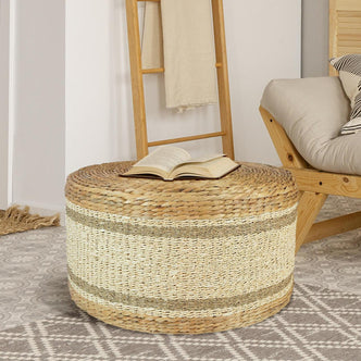 Darcey 75cm Water Hyacinth Ottoman in Natural, used as a coffee table, with an open book on top. Background features a wooden lounge chair and blanket ladder on patterned flooring.