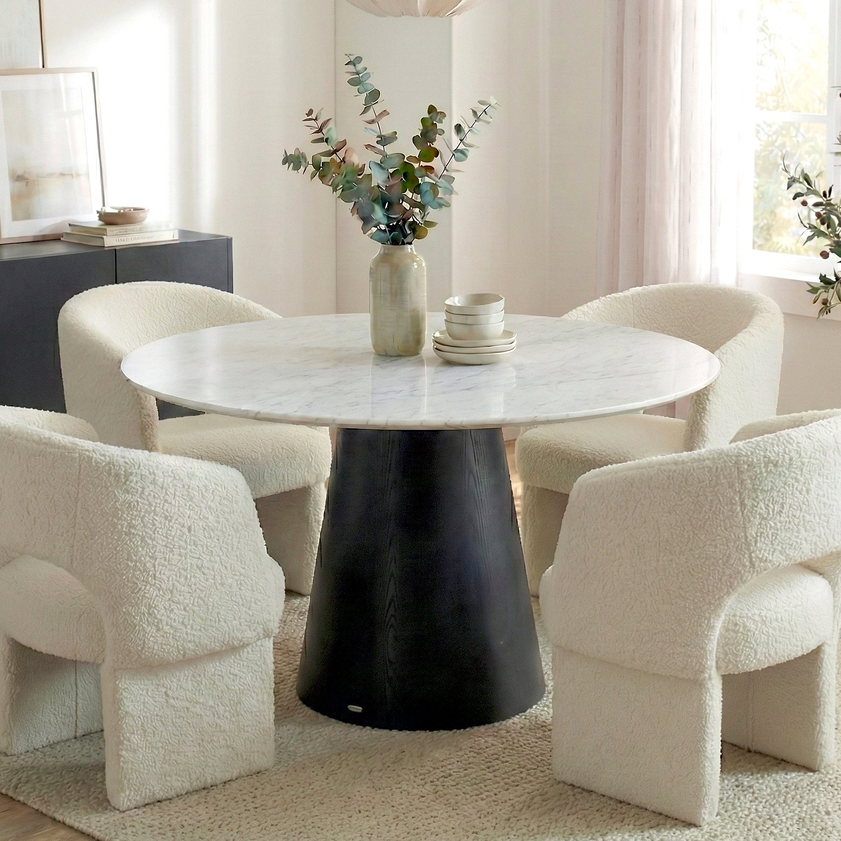Dina 120cm Round Marble Dining Table in Real Carrara Black with white marble top and dark base, surrounded by cream boucle armchairs on a neutral rug, featuring a vase with eucalyptus.