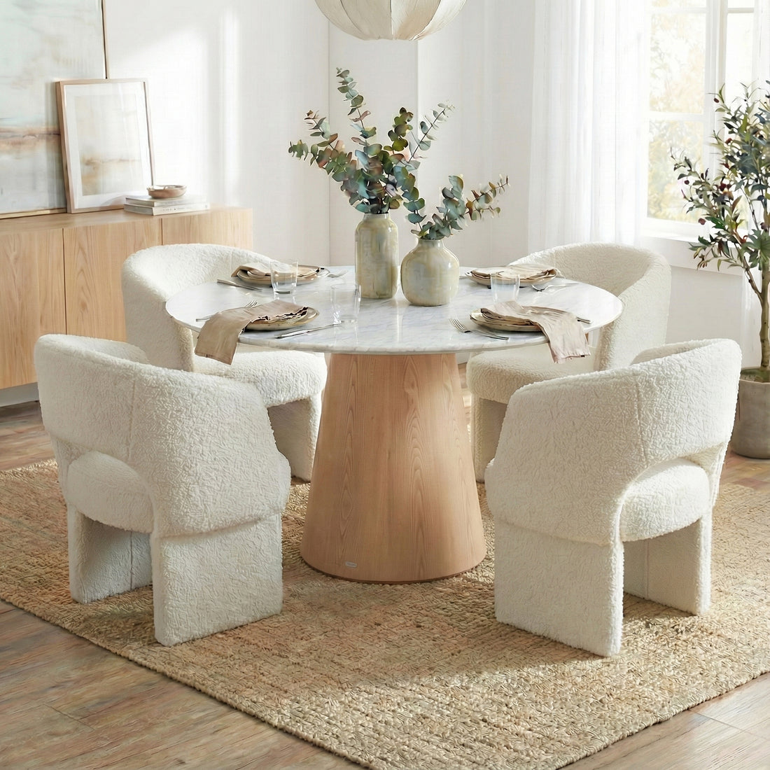 Dina 120cm Round Dining Table in Real Carrara Marble features a marble top with unique veining on a pale wood pedestal, surrounded by plush boucle-upholstered armchairs, set on a jute rug.