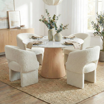 Dina 120cm Round Dining Table in Real Carrara Marble features a marble top with unique veining on a pale wood pedestal, surrounded by plush boucle-upholstered armchairs, set on a jute rug.