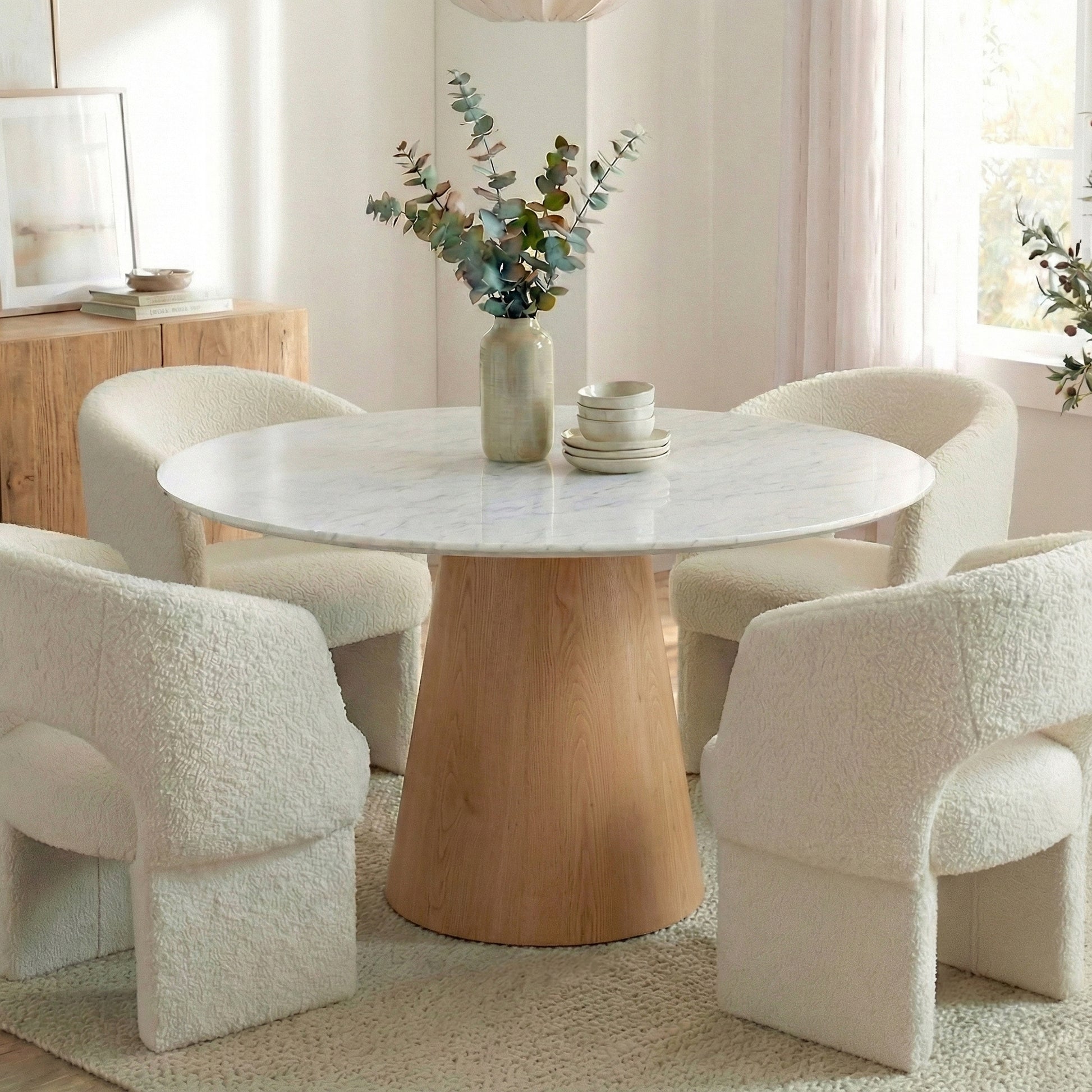 Dina 120cm Round Dining Table in Real Carrara Marble, featuring a marble top with blue-grey veining and a wooden pedestal base, surrounded by boucle-style lounge chairs and a ceramic vase centerpiece.