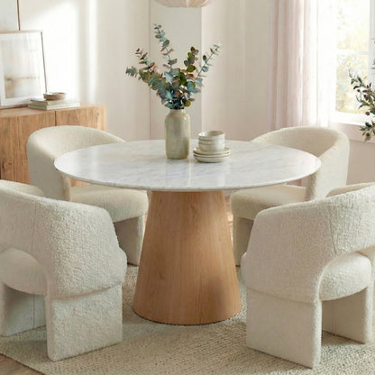 Dina 120cm Round Dining Table in Real Carrara Marble, featuring a marble top with blue-grey veining and a wooden pedestal base, surrounded by boucle-style lounge chairs and a ceramic vase centerpiece.