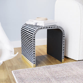 Dustox Mother of Pearl Inlay Side Table in Ivory & Black features a U-shaped profile with black-and-white striped mosaic, brass-tone edges, and surface adorned with books and a wooden bowl.
