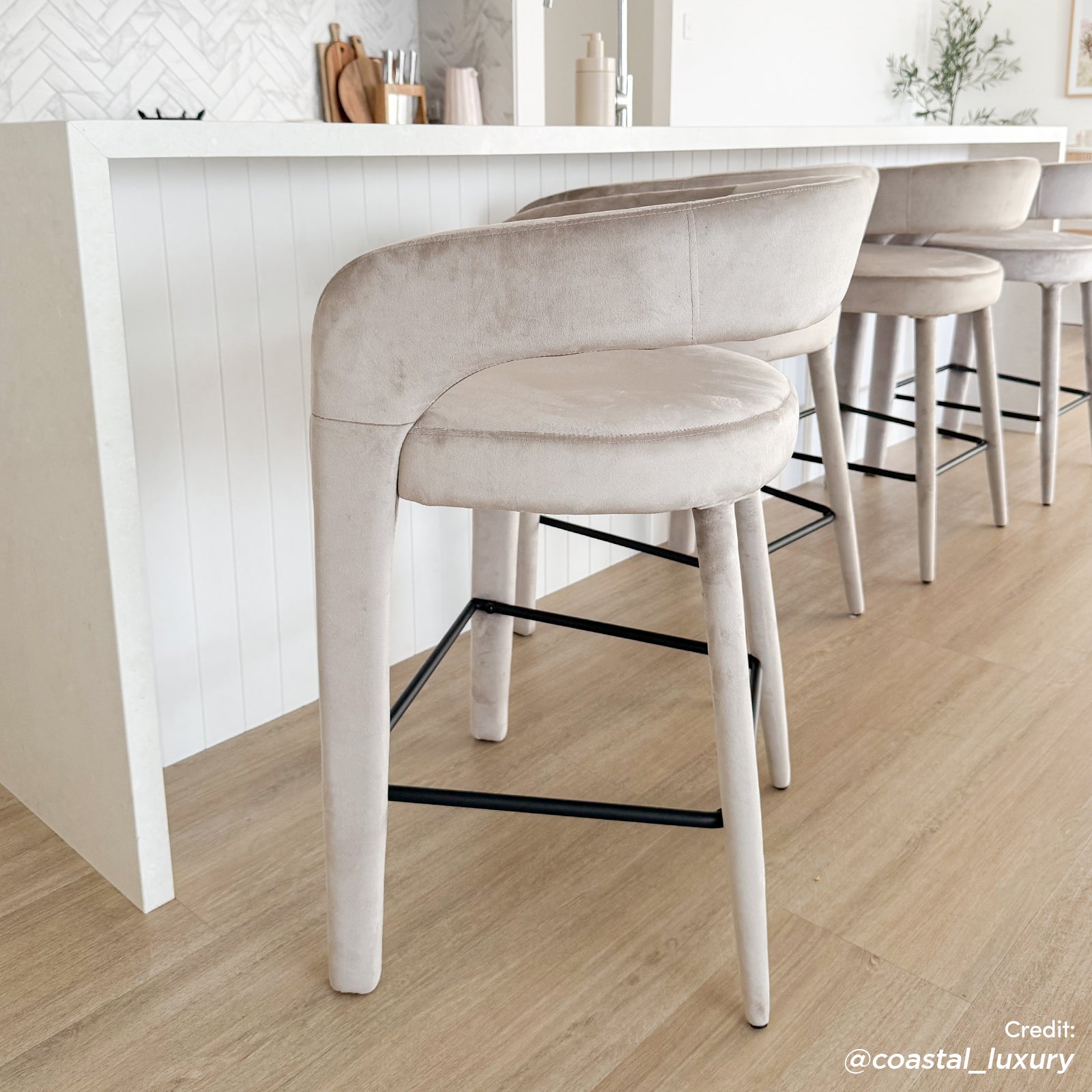 Eisha Velvet Bar Stool Set of 2 in Beige features light-beige, velvet-like upholstery with slender legs and black metal footrests, perfectly complementing the contemporary kitchen island setting.