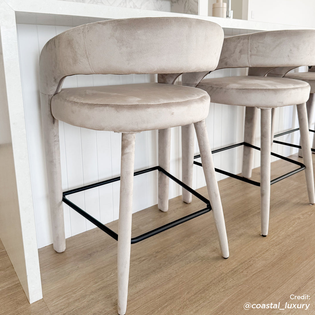 Eisha Velvet Bar Stool Set of 2 in Beige, featuring curved upholstered backrests, round seats, and four fabric-covered legs with a black metal footrest, against a light kitchen island backdrop.