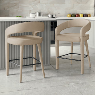 Eisha Velvet Bar Stool Warm Grey Set of 2, featuring a modern design with curved backs and armrests, positioned next to a counter, showcasing their upholstered velvet finish and built-in footrests.