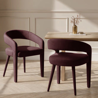 Eisha Velvet Dining Chair Set of 2 in Dark Plum, featuring plush upholstery, low wraparound backrests, and tapered legs, paired with a light wood table on a herringbone floor.