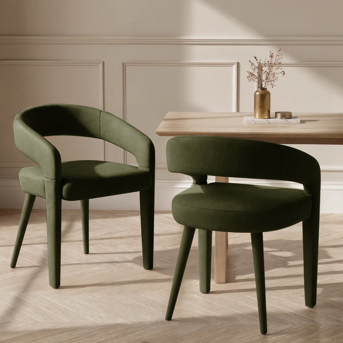 Eisha Velvet Dining Chair Set of 2 in Woodland Green, featuring a rounded open-back design and tapered legs, sits beside a natural-wood table with a brass vase and coasters, on a parquet floor.