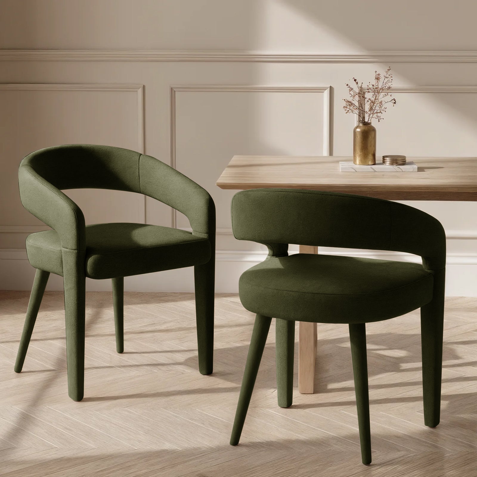 Eisha Velvet Dining Chair Set of 2 in Woodland Green, featuring a rounded open-back design and tapered legs, sits beside a natural-wood table with a brass vase and coasters, on a parquet floor.
