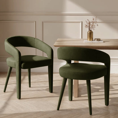 Eisha Velvet Dining Chair Set of 2 in Woodland Green, featuring a rounded open-back design and tapered legs, sits beside a natural-wood table with a brass vase and coasters, on a parquet floor.