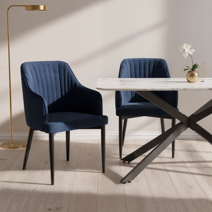 Elly Curved Velvet Dining Chairs Set of 2 in Dark Navy, featuring plush channel-stitched upholstery, slim black legs, and integrated armrests, standing beside a white marble-top table with a black metal base.