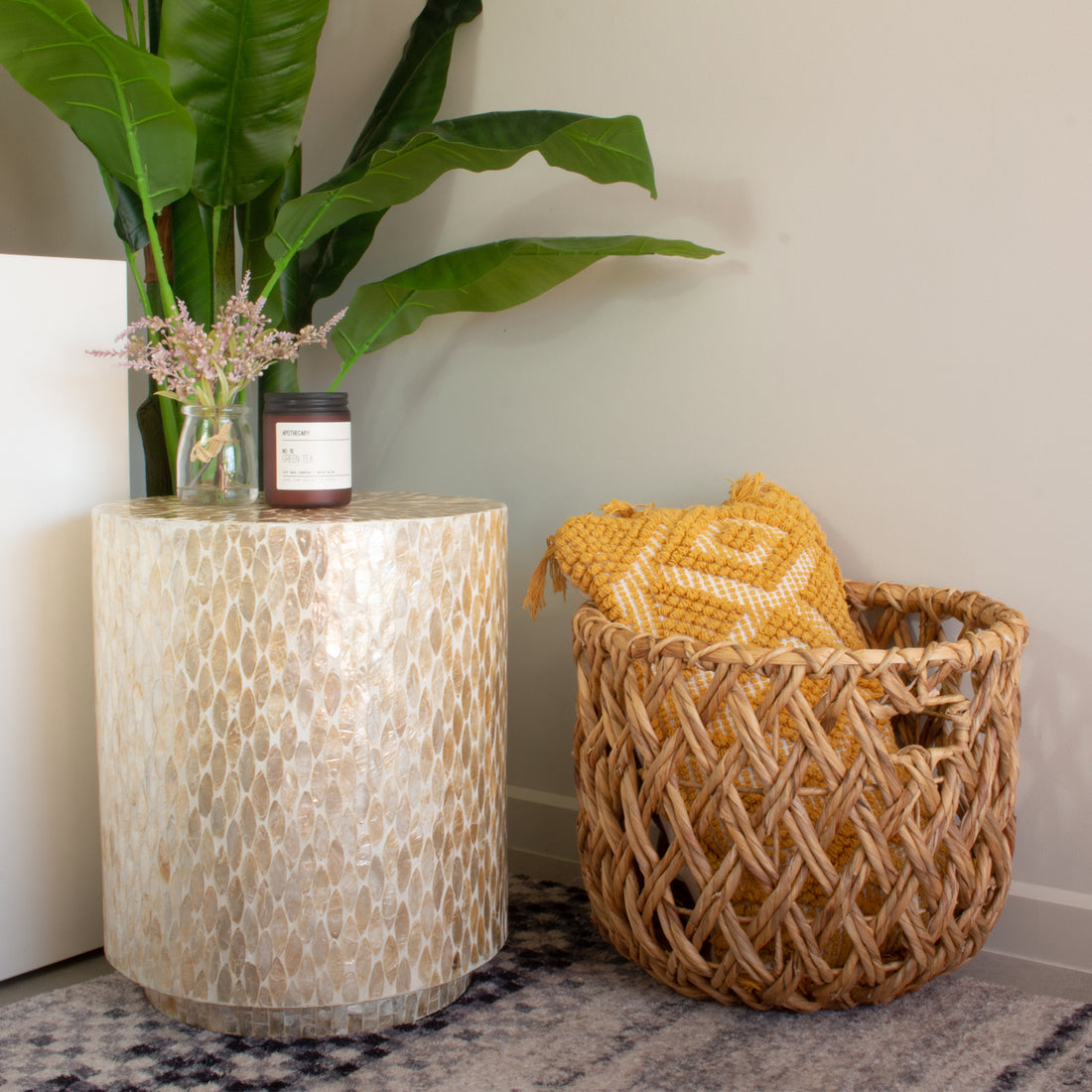 Ely 40cm Mother of Pearl Inlay Side Table in Beige, featuring a stunning mosaic surface, is adorned with a small glass jar of pink blooms and a dark-glass candle, complemented by a mustard-yellow throw in a wicker basket nearby.