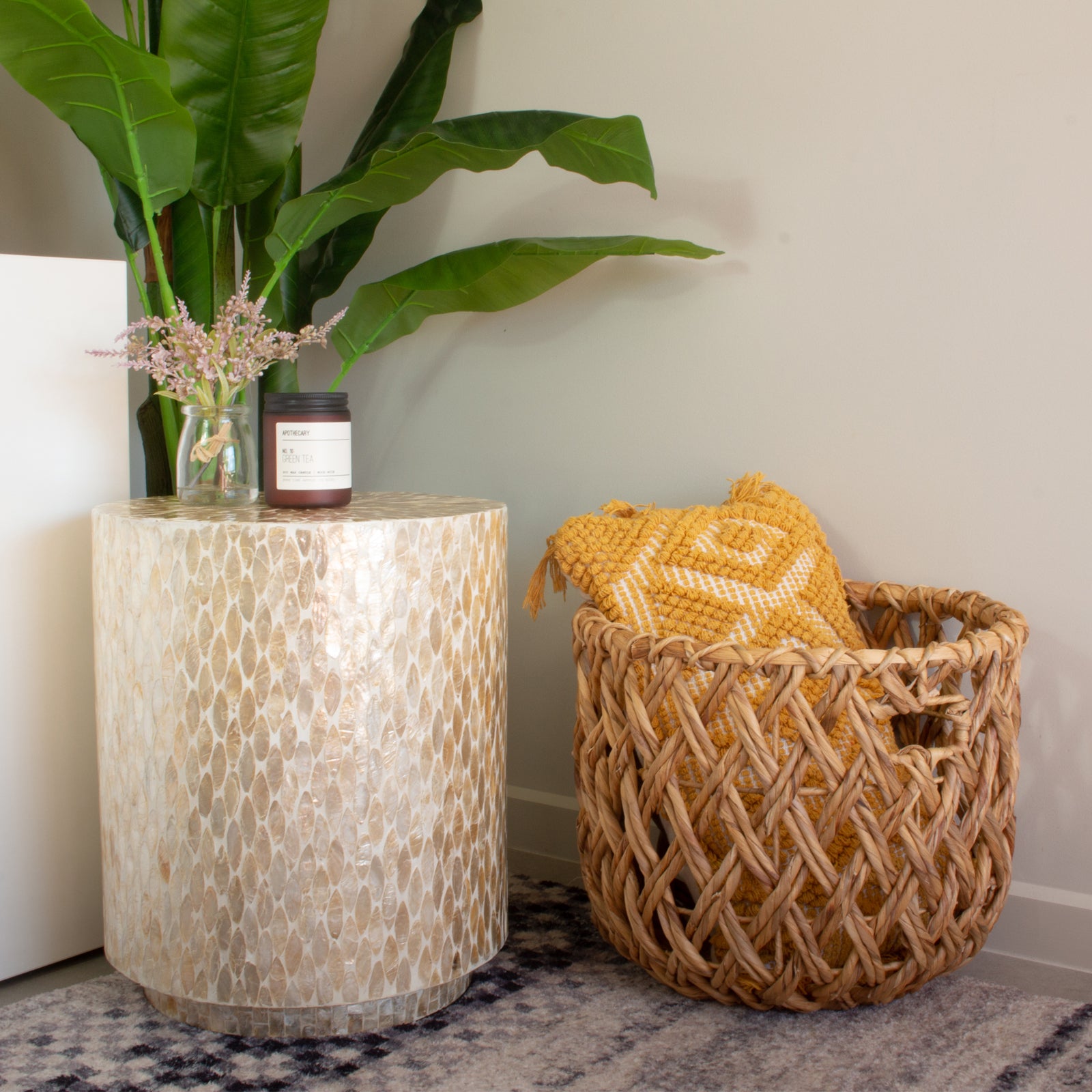 Ely 40cm Mother of Pearl Inlay Side Table in Beige, featuring a stunning mosaic surface, is adorned with a small glass jar of pink blooms and a dark-glass candle, complemented by a mustard-yellow throw in a wicker basket nearby.
